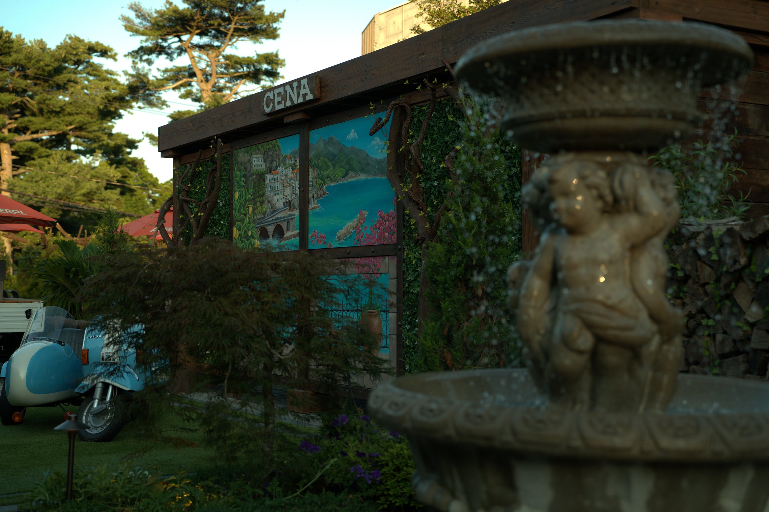 A garden scene featuring a statue in the foreground, a mural with the word 'CENA' and a lakeside landscape, trees, and umbrellas in the background.
